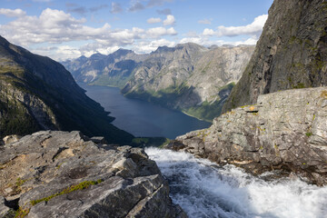 View of the landscape with the source of the Mardalsfossen waterfall in Norway. Grey sky with clouds, grey mossy rocks, white water of the waterfall and dark blue Eikesdalsvatnet lake