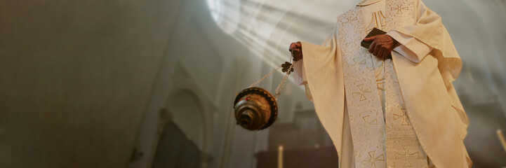 Website banner with unrecognizable priest holding Bible and majestic golden censer while besmoking...