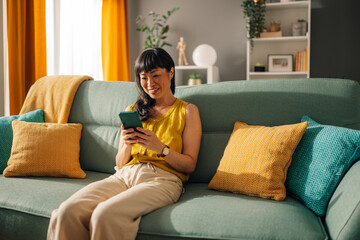 Asian woman smiling while using smartphone on green couch at home