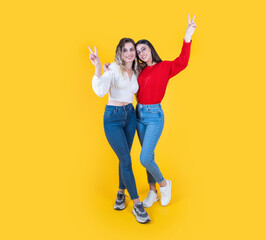 Full body happy two 20s caucasian women besties posing together showing v sign smiling over yellow studio background with copy space. Wear casual shirts and jean. Lifestyle concept idea image.