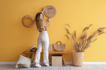 Young woman hanging mirror on yellow wall in hallway