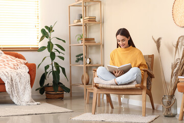 Young woman sitting in armchair and reading book in living room