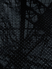 Close-up of raindrops creating ripples on a dark, wet metal surface with reflective shadows of a fence and trees.