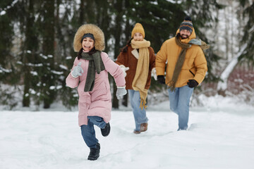 Full length portrait of carefree young girl running towards camera in winter forest with family in background and enjoying holiday vacation together copy space