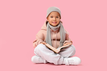 Cute little Asian boy in warm clothes with book sitting on pink background