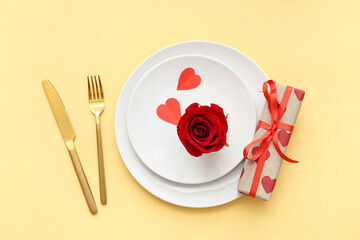 Beautiful table setting with red rose, paper hearts and gift box on yellow background. Valentine's Day celebration