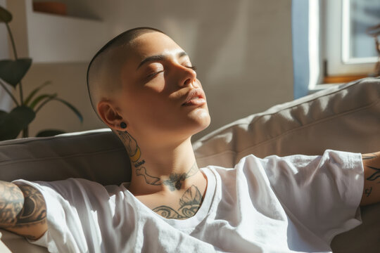 Non-binary individual with tattoos relaxing on a couch, eyes closed and basking in warm sunlight, expressing a peaceful and contemplative mood
