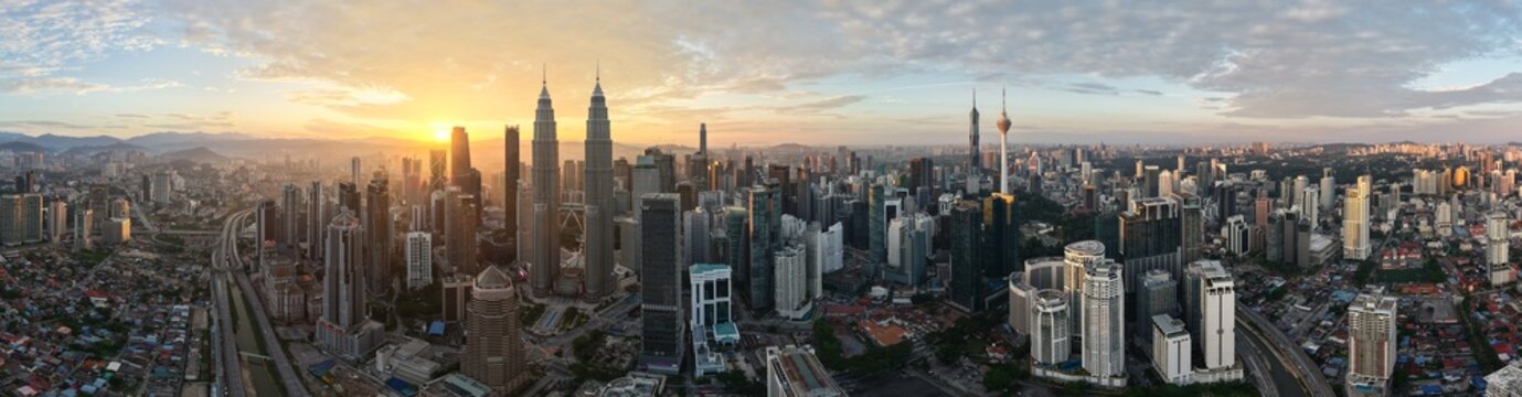 Wide aerial panorama view of Kuala Lumpur skyline with sun shining from behind buildings. Drone pano of KLCC including view of Petronas Towers, The Exchange 106, KL Tower and Merdeka 118.
