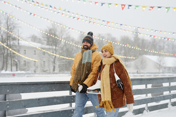 Waist up portrait of happy young couple holding hands walking by skating rink in winter park and...