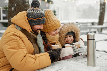 Portrait of modern young family drinking hot cocoa outdoors in winter sitting at cafe table together and smiling happily