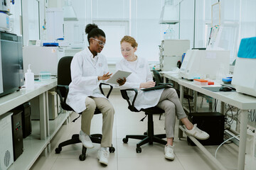 Two scientists sitting in laboratory, analyzing data on tablets while discussing research. Laboratory equipment and samples placed on counters around them