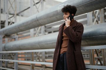 Fashionable man in brown coat talking on cell phone in city, standing in front of metal railing on...