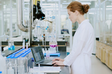 Scientist working in laboratory wearing white lab coat, using laptop for research. Laboratory setting with various scientific equipment and glassware visible in background