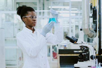 Black scientist analyzing a blue liquid sample in modern laboratory environment, wearing white lab...