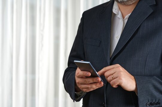 Confident businessman in a tailored suit actively using his smartphone for communication while standing in a spacious, modern office, showcasing professionalism and focus on work.
