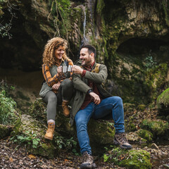 husband and wife hikers sit in front cave and drink from metal mug