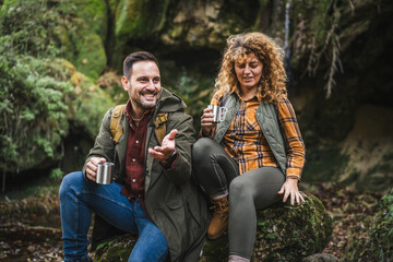 husband and wife hikers sit in front cave and drink from metal mug