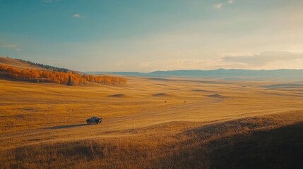 Obraz premium A lone car drives through a vast, golden field with a line of trees in the background as the sun sets over the horizon.