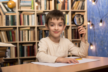 Close-up of a child holding a magnifying glass at a table with colored pencils and a blank sheet of...