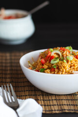 A white bowl filled with rice and tender meat, garnished with fresh tomato chunks, sliced scallions and parsley. The meal sits on a bamboo placemat, with a blurred sauce container in the background