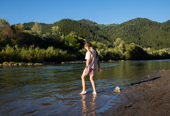 Fototapeta premium A girl stands in shallow water, feeling the cool river flow around her feet while sunlight filters through surrounding trees, creating a serene atmosphere filled with nature's beauty.