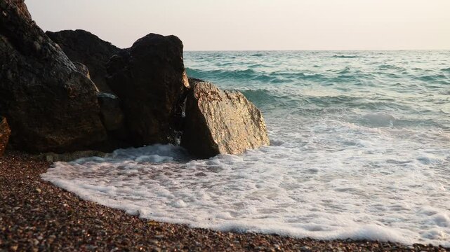 Waves gently lap at rocky shore during stunning sunset over ocean. Golden sunlight reflects off waves as they roll onto shore, creating tranquil atmosphere at sunset by sea. Montenegro summer, Canj