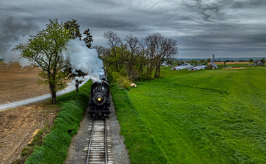 A classic steam locomotive steams along the tracks surrounded by vibrant green fields and trees, passing near a quaint rural farm under a dramatic cloudy sky.