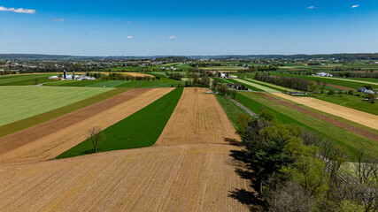 A patchwork of green, golden, and brown fields stretches across the landscape, showcasing cultivated farmland under a bright blue sky. Distant farms and trees dot the horizon.