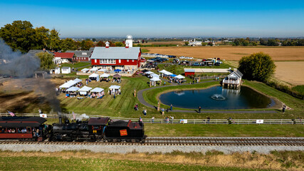 A steam train travels along the tracks near a bustling rural fair, with families gathered around tents, enjoying activities. A serene pond and farm buildings are visible under a bright blue sky.