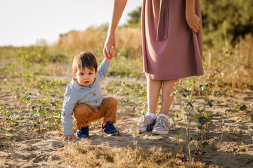 Mom holds her one-year-old baby by the hand for a walk in the park by the river