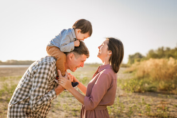 Happy family walking on sandy beach of river. Father, mother holding baby son on hands and going together. Rear view. Family Ties concept.