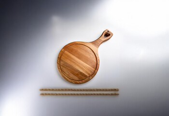 A round wooden cutting board with a handle sits on a white surface with two sticks of wood beside it.