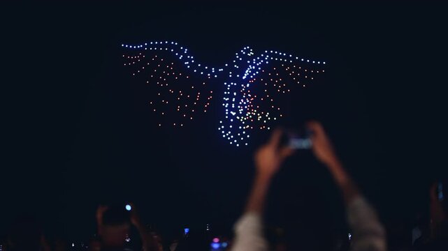 illuminated aerial drone show at glowing night sky, programmed flying robotics light performance over city, people watching and filming, advanced technology concept