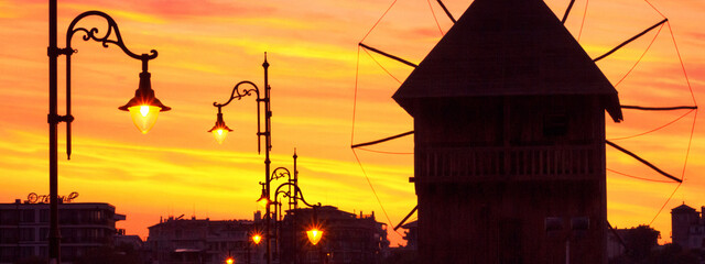 Evening city landscape, banner - view of the silhouettes of the wooden windmill and street lamps on the embankment of Nessebar before the entering to the Old Town, on the Black Sea coast of Bulgaria