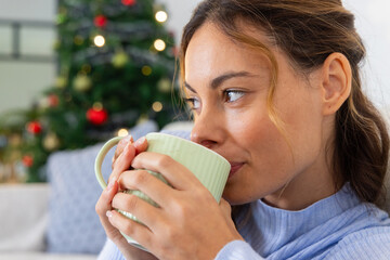 Woman enjoying hot drink at home near Christmas tree, feeling cozy and relaxed