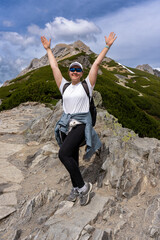 Happy female hiker with arms raised stands on a rocky mountain trail with a scenic view. Tourism, active lifestyle, mountain travelling, hiking.