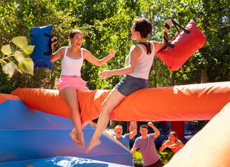 Emotional happy young girl funny pillow fighting on inflatable log with female friend in outdoor...