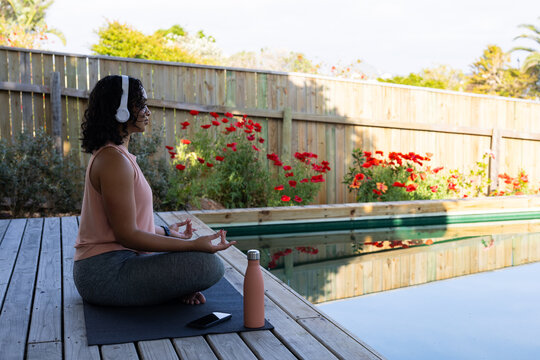 Woman meditating by pool, wearing headphones, enjoying peaceful outdoor setting