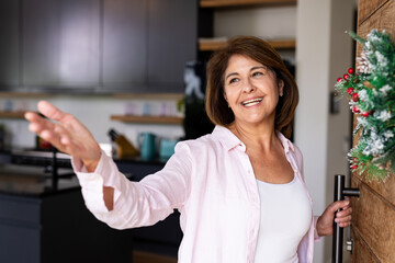 Christmas time, Welcoming guests, smiling senior woman opening door with festive wreath at home