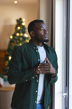 African american man standing by window, contemplating holiday season with Christmas tree glowing