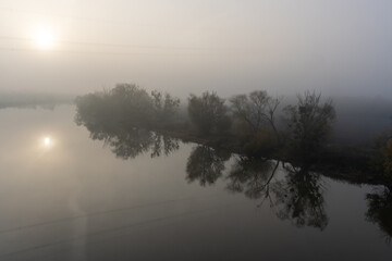 Fototapeta premium Serene misty sunrise over calm river with soft reflections of trees and gentle, hazy sky. Concept of peaceful nature, early morning tranquility, and foggy landscapes.