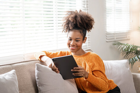 Happy young black woman using digital tablet relaxing on sofa at home.