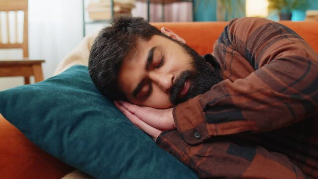 Tired Indian young man lying down in bed taking a rest. Carefree peaceful guy napping, falling asleep on comfortable sofa with pillows at home living room. Closes his eyes enjoy daytime nap alone