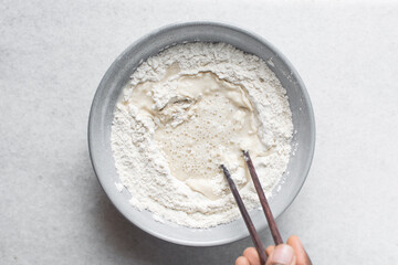 Overhead view of noodle dough being being mixed in a grey bowl, top view of flour and water being mixed with chopsticks to make noodles, process of making hand pulled noodles