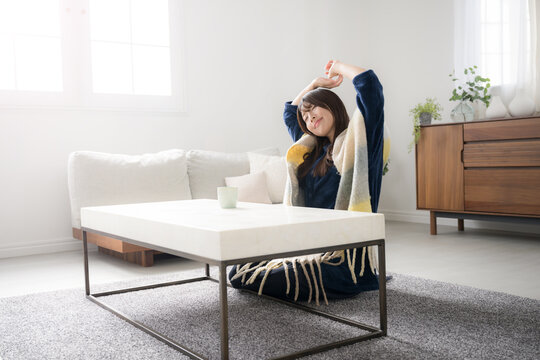 Young woman in pajamas sitting and stretching in her living room Relaxing on a late waking fall/winter morning Wide angle