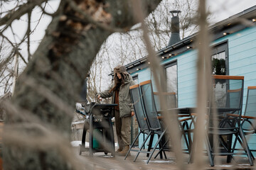 stylish girl in a warm jacket and hat in the yard of a tourist house is grilling meat and drinking coffee, enjoying a rural vacation and natural scenery