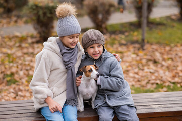 Brother and sister sit in an embrace with a dog on a bench for a walk in the autumn park. Boy, girl and jack russell terrier. 