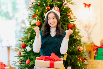 Young Asian woman wearing a red and white Christmas hat surrounded by gift boxes and holding a red...