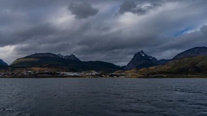 The picturesque mountain range of the Andes. Snow-capped peaks against the sky and clouds. At the foot, on the ocean shore, the city of Ushuaia is visible. In the foreground is the water surface 