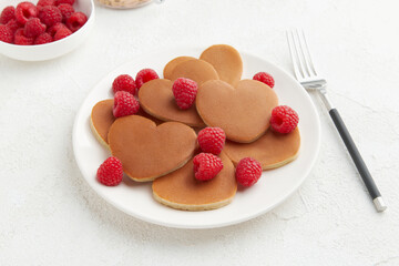 A heap of heart shaped pancakes with raspberries and a fork on the white plate on white background